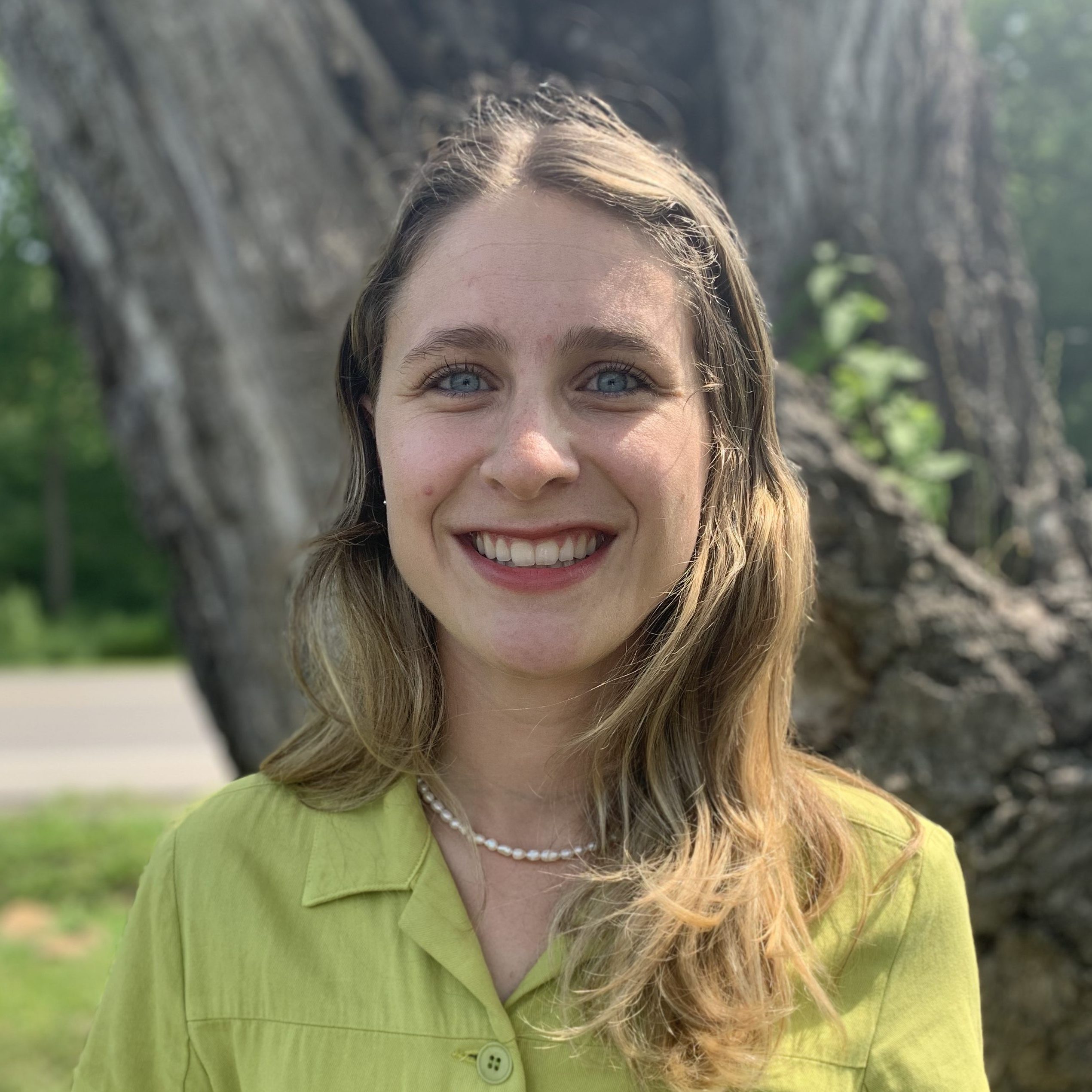 A portrait of a woman with long light hair, wearing a beaded necklace and light blouse, smiling.